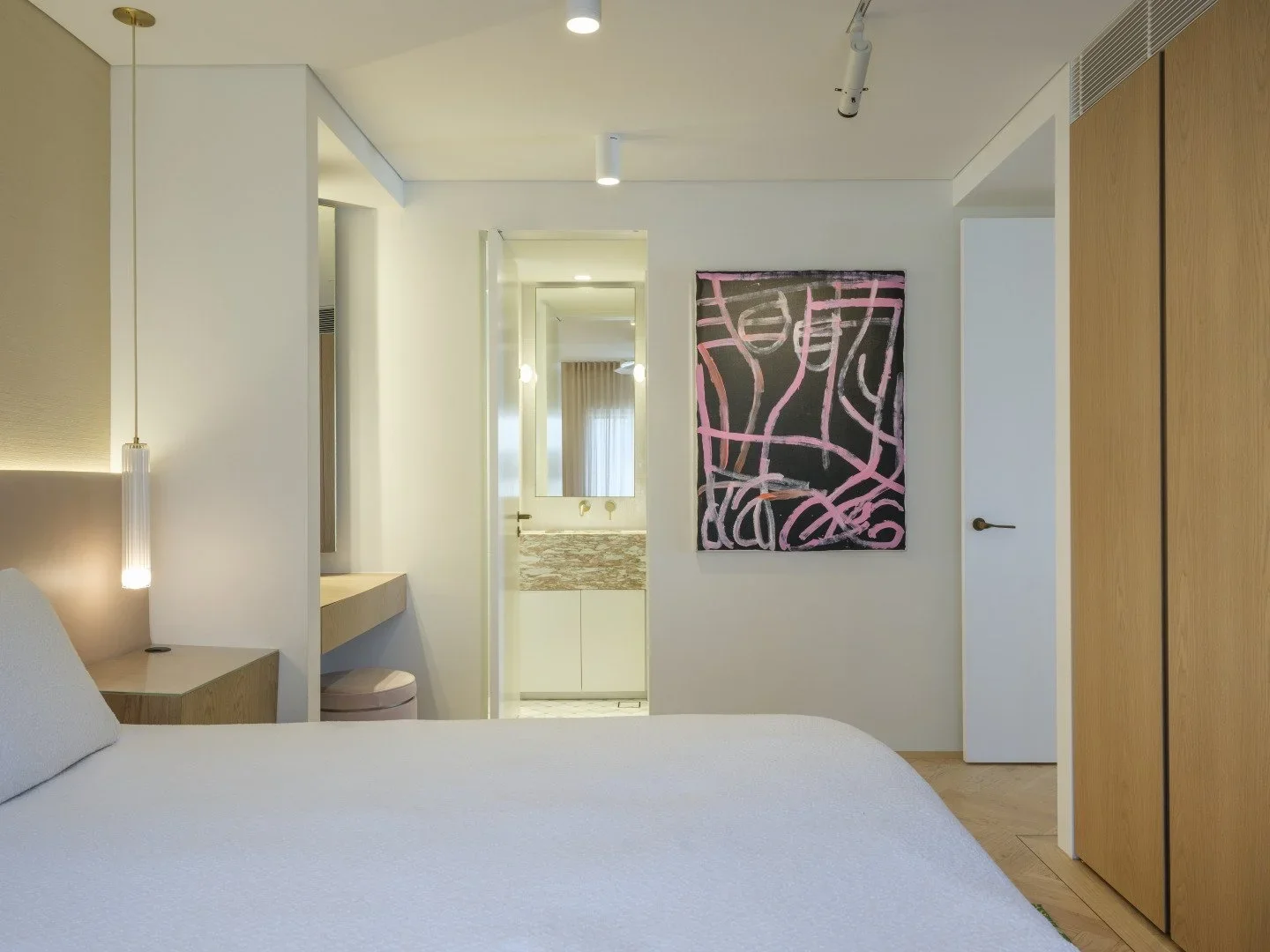 Modern hotel room with white walls, beige bedding, and a wooden wardrobe. A nightstand with a hanging pendant light is visible on the left. A colorful abstract painting is on the wall, and a doorway to a bathroom with a marble countertop and a mirror