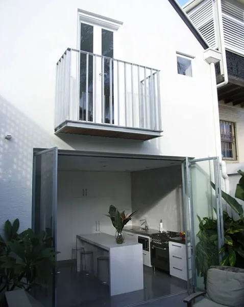 Modern outdoor kitchen with open glass doors, white countertops, and a kitchen stove under a small balcony on a white house exterior