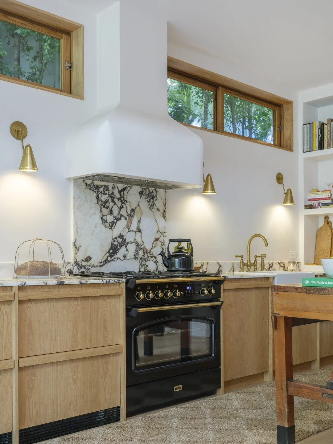 Kitchen with wooden cabinets, a black stove with gold knobs, a marble backsplash, a kettle on the stove, brass wall-mounted lights, a window with greenery outside, a white countertop, and books on a built-in shelf.