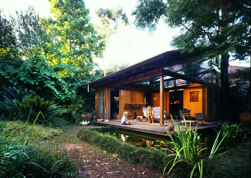 A modern house with large glass windows and a patio, surrounded by greenery, with two children and an adult sitting and talking on the patio.