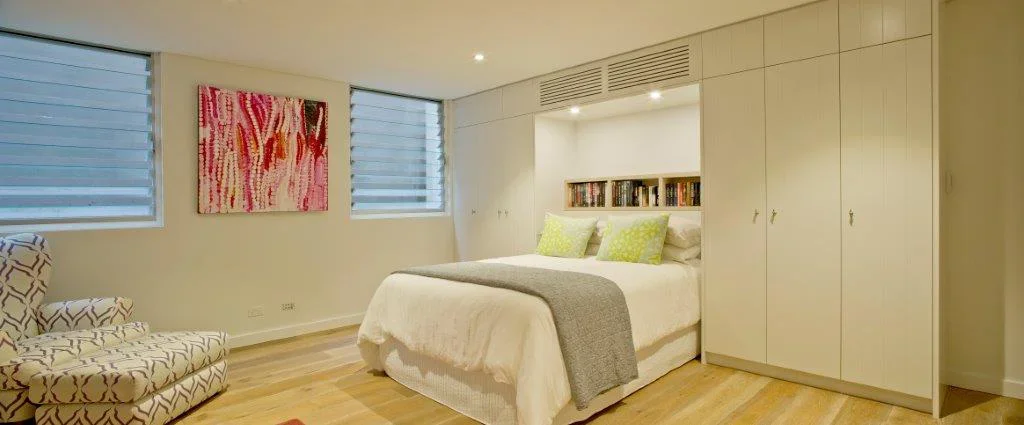 A modern bedroom with a bed, two green pillows, built-in wardrobe, and a wall-mounted bookshelf above the bed, with two windows with blue blinds and a patterned armchair in the corner.