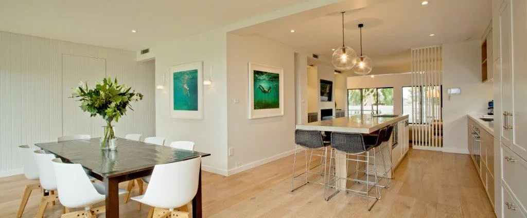 Open-concept kitchen and dining area with white walls, wooden flooring, a dining table with white chairs and a large floral centerpiece, a kitchen island with bar stools, and sliding glass doors leading outside. Artworks and modern light fixtures are