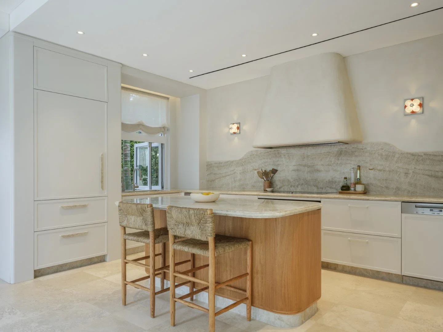 Modern kitchen with white cabinets, a marble island, wooden chairs, and a window with a view of greenery.