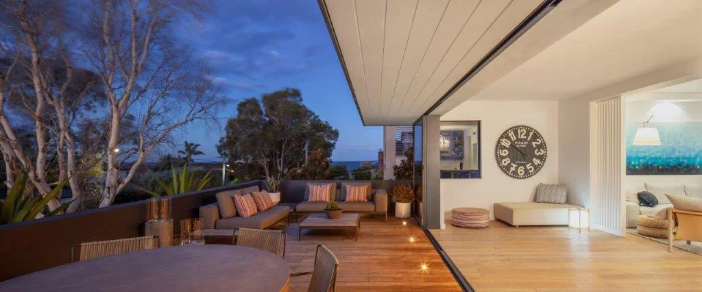 Outdoor balcony with seating area and view of trees at dusk, adjacent to an indoor living space with wall clock and artwork.