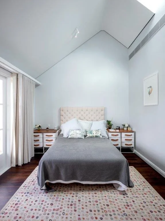 A bedroom with a bed, two nightstands, and a large window with curtains, in a room with angled ceiling and minimal decor.