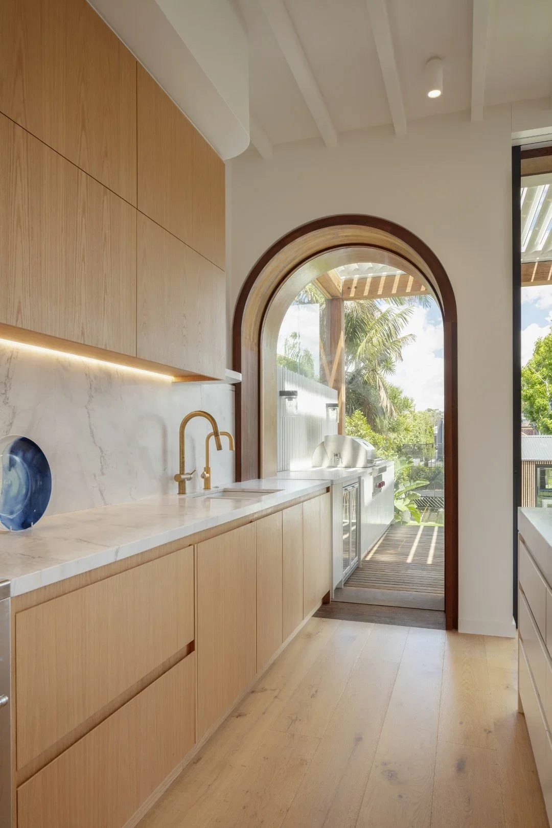 Modern kitchen with wooden cabinets, marble countertop, gold faucet, and an arched window with view of outdoor garden and patio.