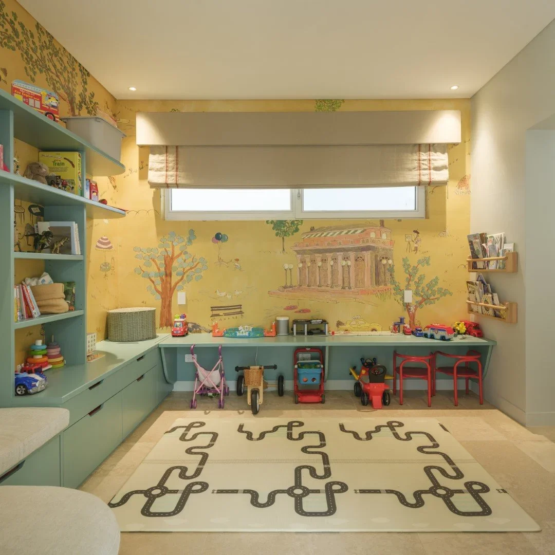 Colorful children's playroom with a mural of painted trees and classical building on yellow wall, toy cars, bikes, a stroller, a bookshelf, and chairs, with a beige and black patterned area rug on the floor.