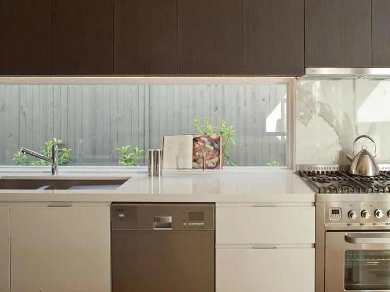 Modern kitchen with white countertops, a built-in dishwasher, a stainless steel stove with a kettle, and a window with a view of green plants outside.