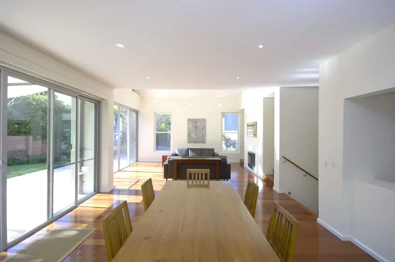 Bright open living and dining area with large sliding glass doors, wooden flooring, a wooden dining table with six chairs, and a gray sofa with a wall-mounted artwork in the background.