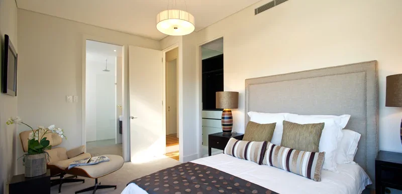 A modern bedroom with a large bed, beige upholstered headboard, white bedding, and decorative pillows. There are two side tables with lamps, a small sitting area with a chair and flowers, and an open door leading to a closet or bathroom. The room is 