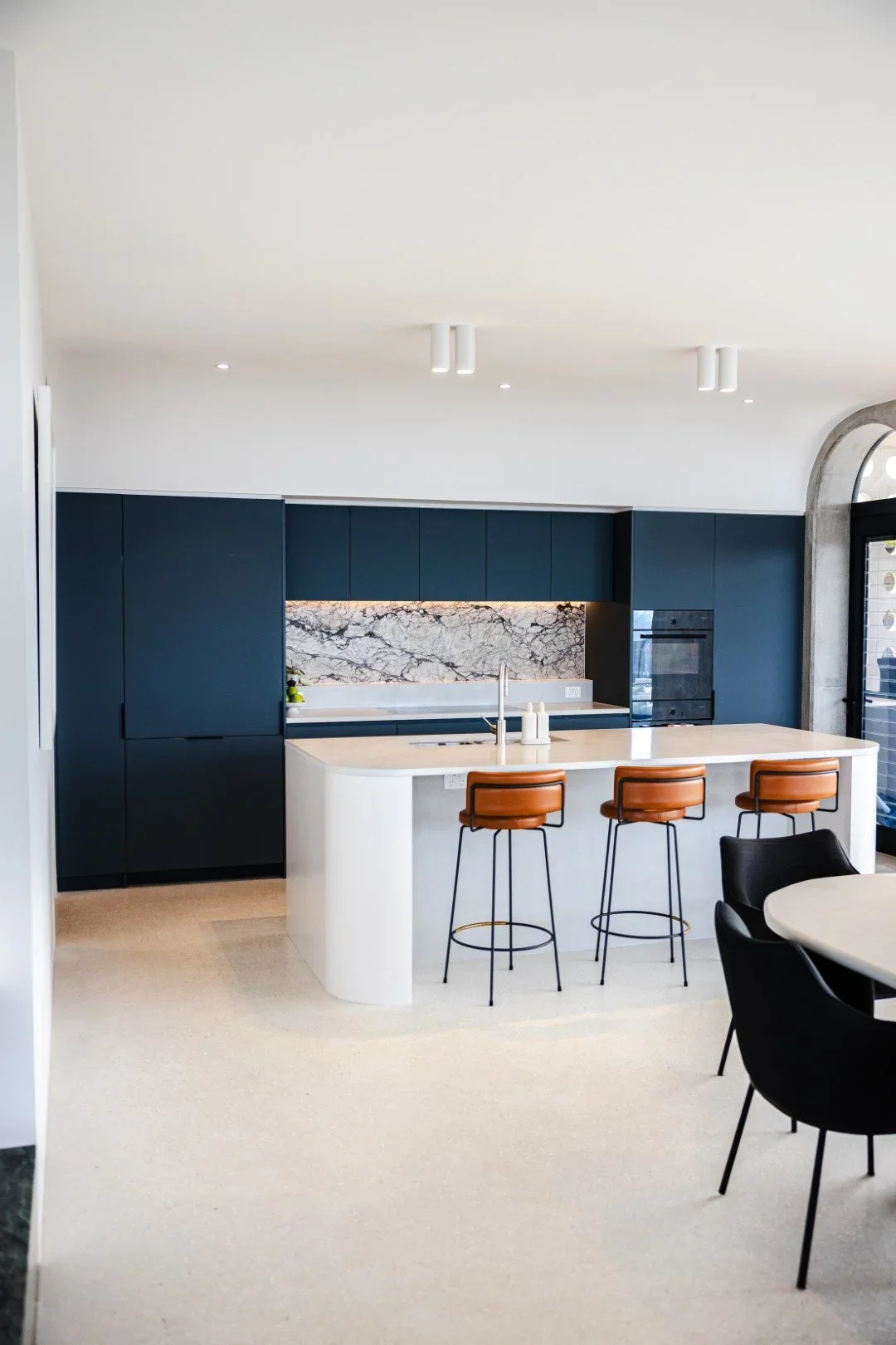 Modern kitchen with navy blue cabinets, marble backsplash, white island with tan bar stools, and black chairs around a dining table.