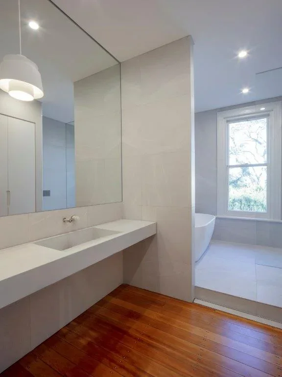 Modern minimalist bathroom with a white vanity, large mirror, wood floor, and a separate shower area with a bathtub near a window.