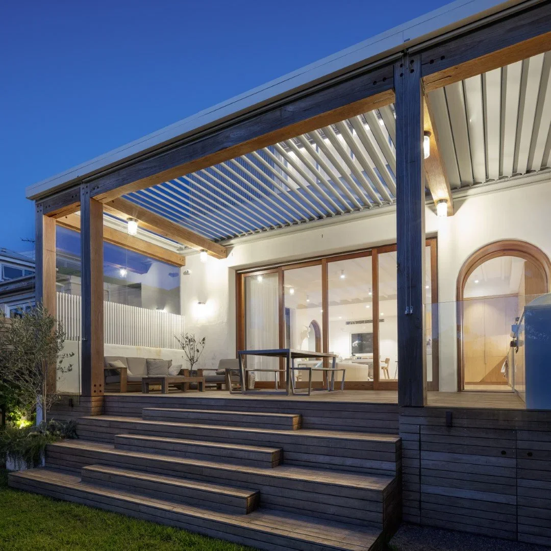 Exterior view of a modern house patio with wooden steps leading up to a covered seating area, illuminated by outdoor lights at dusk.