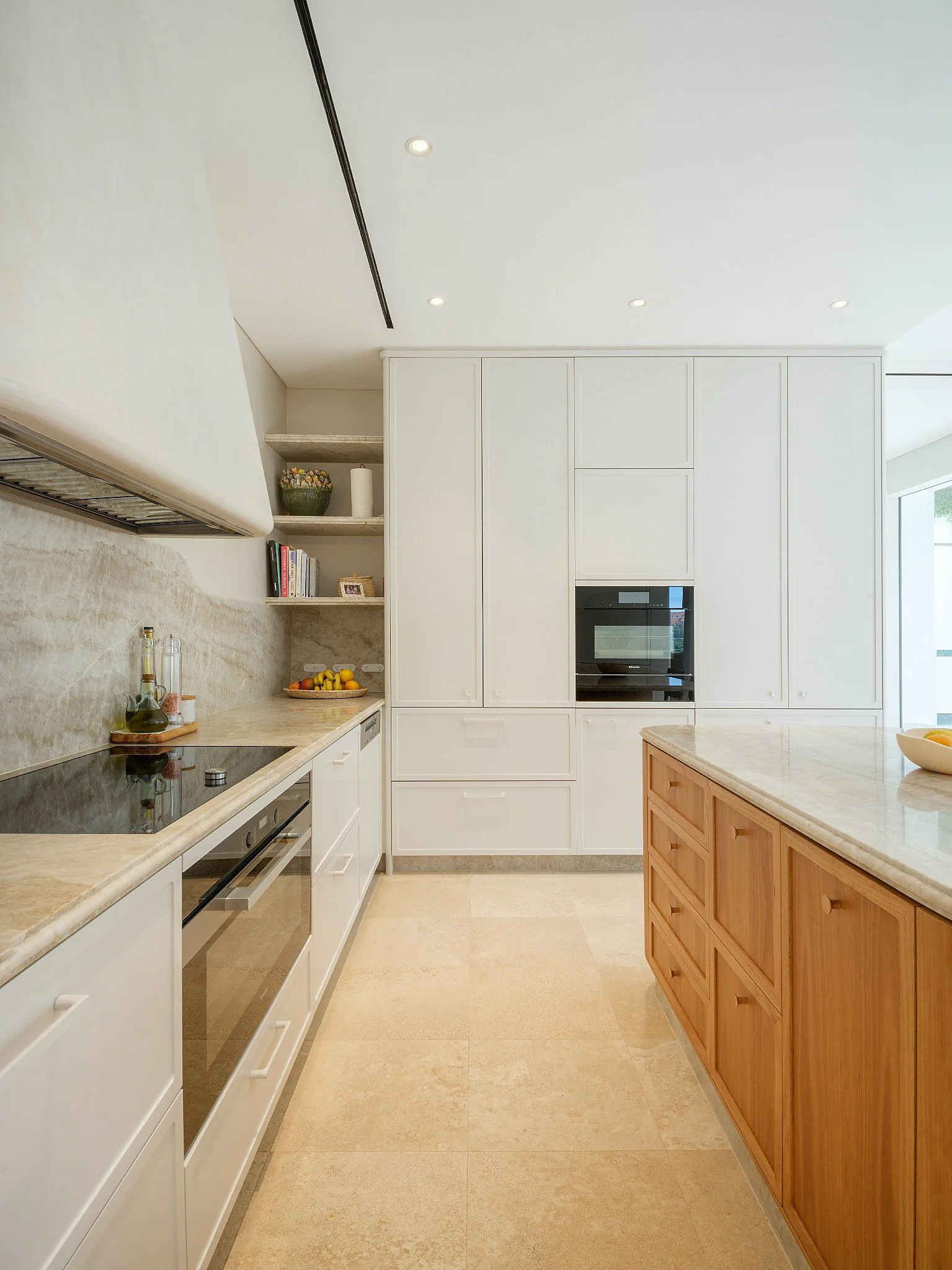 Modern kitchen with white cabinets, marble island, two beige bar stools, grey textured backsplash, and built-in appliances, illuminated by ceiling and wall lights.