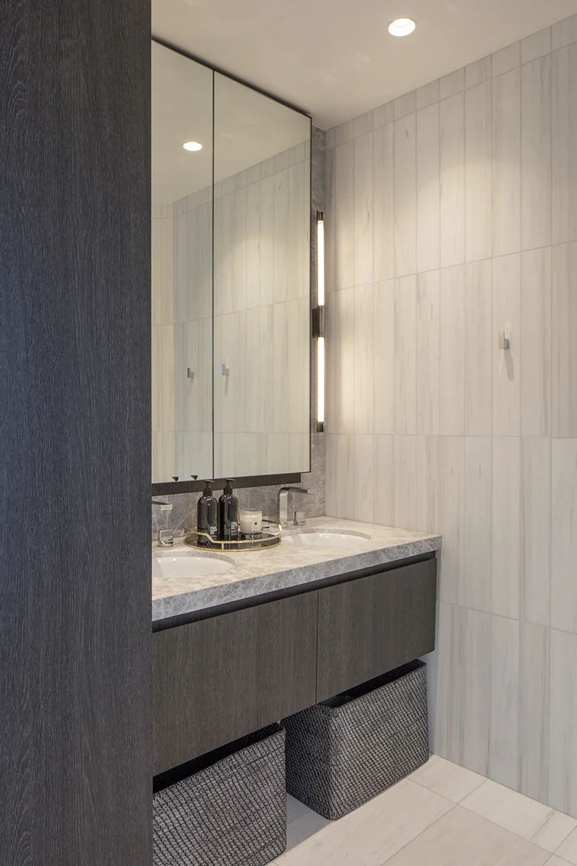 Modern bathroom with a large mirror, double sink vanity, and sleek lighting fixture on a marble wall.