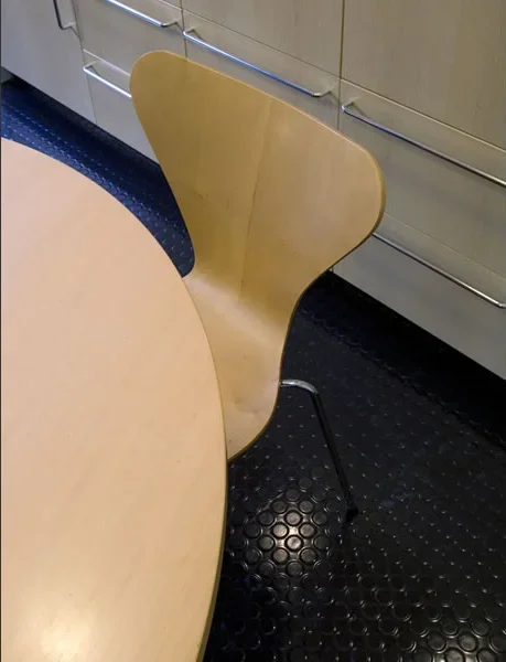 Wooden chair with a curved backrest and metal legs next to a light-colored round table, in a room with a black textured floor and light storage cabinets.