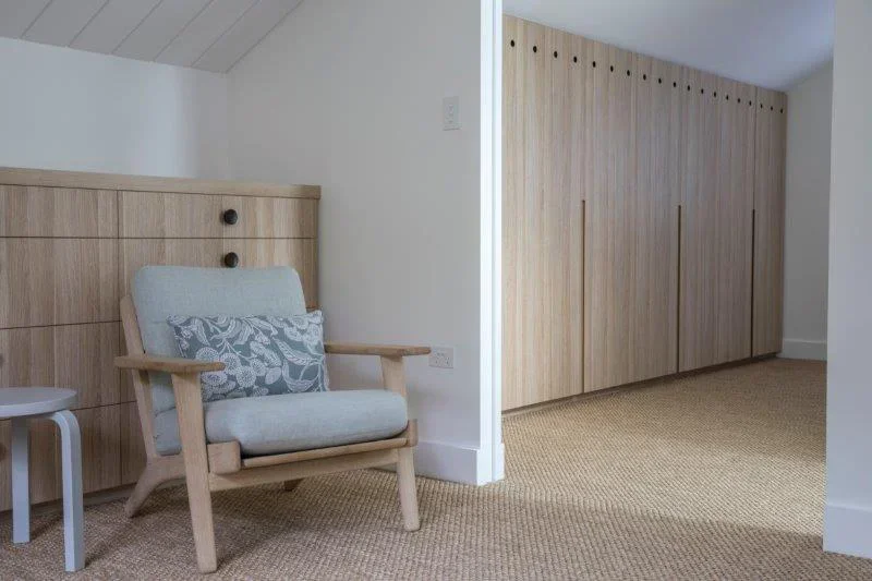 A light-colored wooden armchair with light blue cushion and patterned pillow, next to a small white side table, in a room with beige carpet and built-in wooden wardrobe doors.