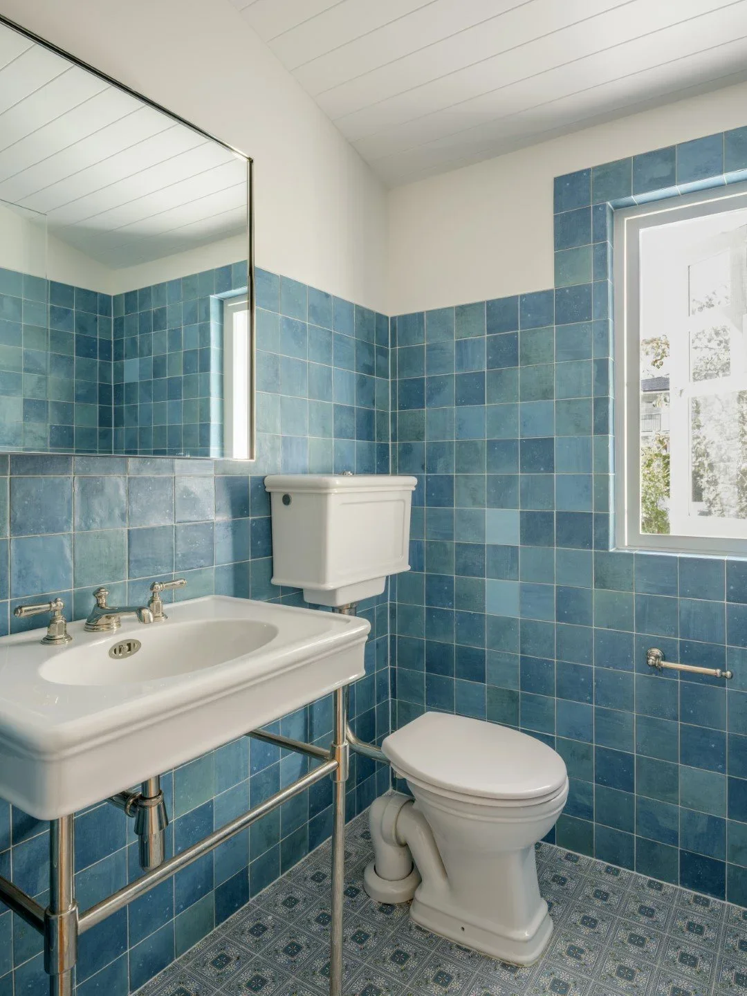 Small bathroom with blue tiled walls, a white porcelain toilet, white porcelain sink with metal supports and faucets, a large mirror above the sink, a window letting in natural light, and patterned tile flooring.