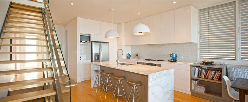 Modern open-concept kitchen with white cabinets, marble island, stainless steel refrigerator, and wooden bar stools.