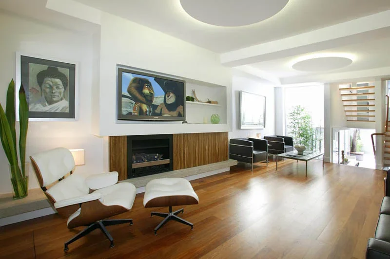 Modern living room with white walls, hardwood floors, a wall-mounted flat-screen TV, black armchairs, a white lounge chair with matching ottoman, framed artwork, large windows with sheer curtains, and decorative plants.