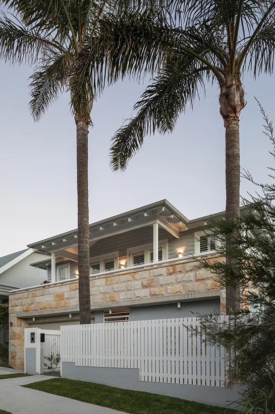 A modern house with a stone facade, white balcony railing, and exterior lighting, framed by tall palm trees and a white picket fence.