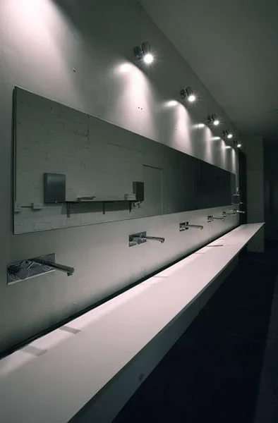 Empty bathroom sinks with faucets, mirror, and wall-mounted fixtures in a modern, minimally lit space.