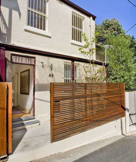 View of a two-story white brick house with an open front door, a wooden slatted fence, and greenery in the background.