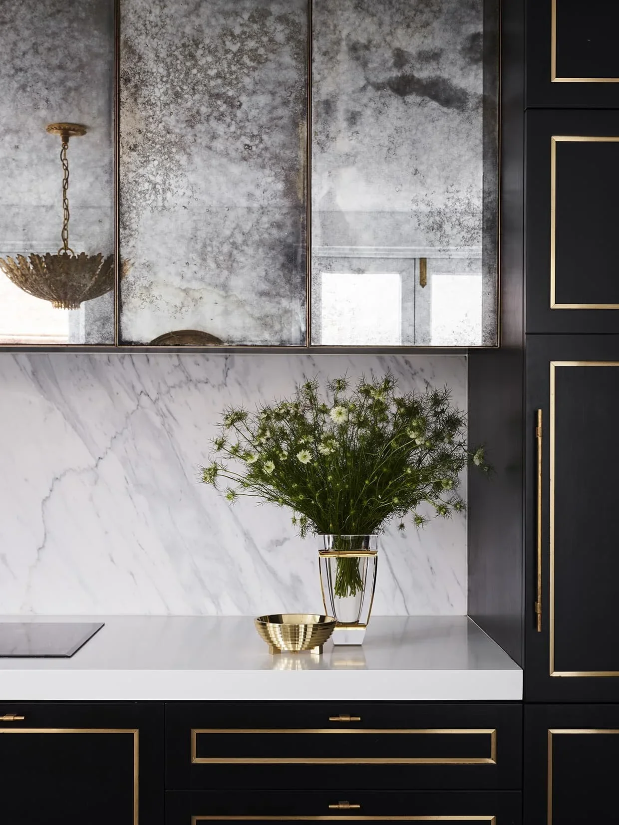 Modern kitchen with black cabinetry with gold trim, marble backsplash, glass vase with green flowers, and reflective cabinet doors.