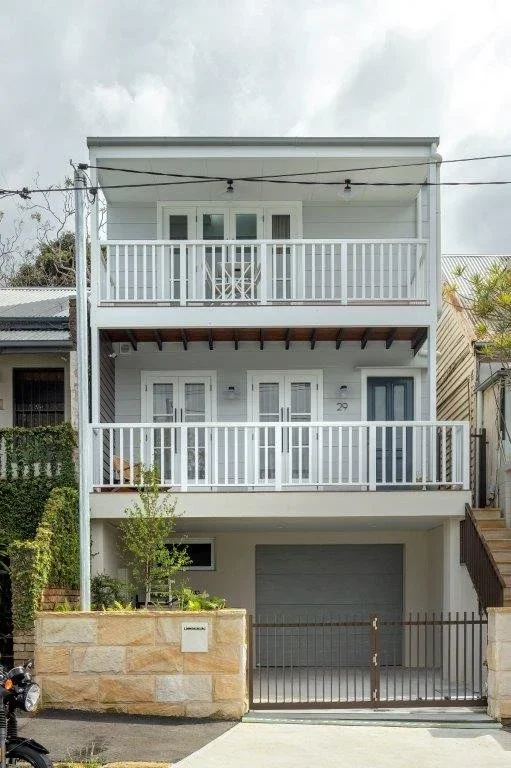 Three-story white house with balconies on the second and third floors, a garage on the ground level, and a small front yard with a tree. Gated entrance to the garage.