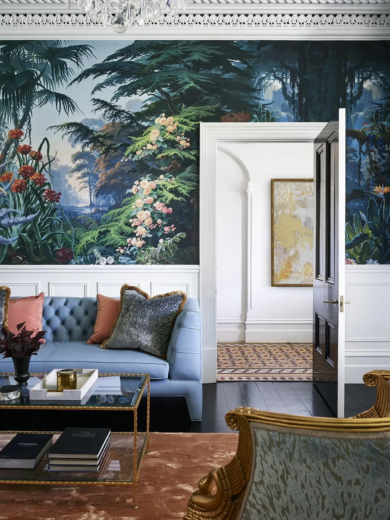 A luxurious living room features a large, colorful mural of a lush, green forest scene on the wall behind a light blue tufted sofa. The sofa is adorned with pastel pink and gray pillows. In front of the sofa, there's a glass coffee table with a gold 