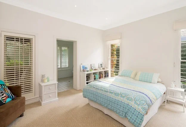 Bright bedroom with white walls, a large bed with a light blue and white quilt, and windows with white blinds, a bookshelf, a brown armchair, and other small furniture.