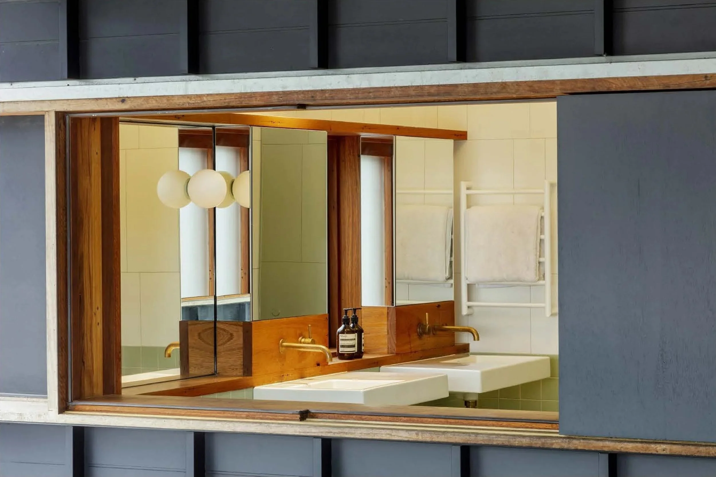 Interior view of a modern bathroom with two white sinks, wooden mirror frame, black soap dispensers, and a towel rack with towels on the wall.