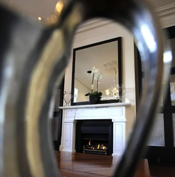 Reflection of a white fireplace with a black insert, a large mirror, a potted plant with white flowers, and decorative glassware on the mantel, seen through a blurred gold and black decorative object.