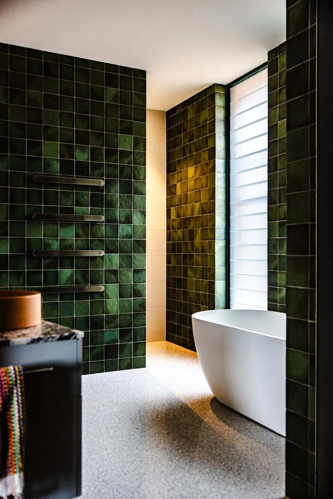 Modern bathroom with dark green tiled walls, a large window with white blinds, and a sleek white freestanding bathtub.