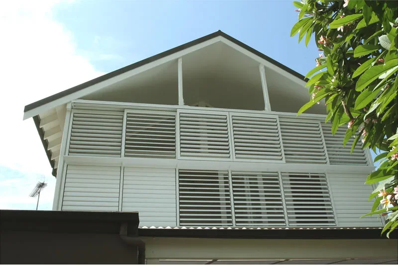 A two-story house with white horizontal slat window shutters on the upper floor, a sloped roof, and a tree with green leaves partially in the frame on the right side.