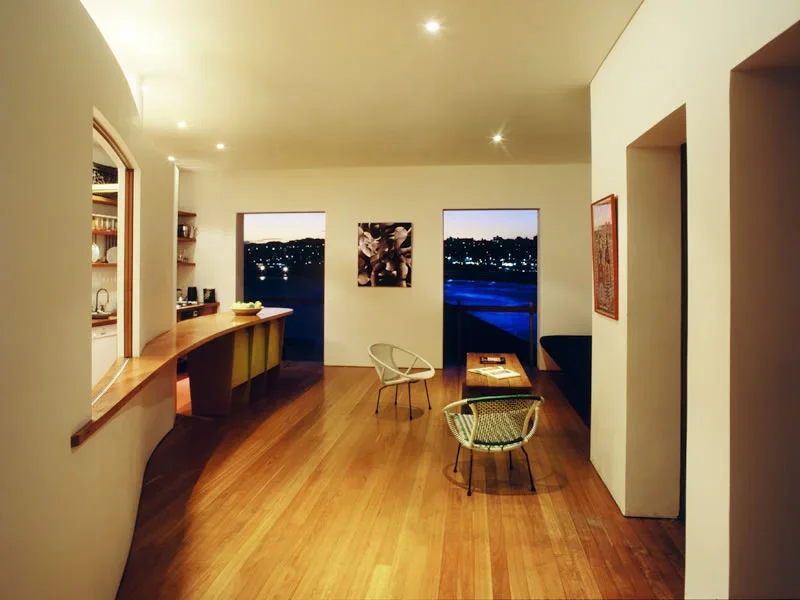 Living room with wood floors, two large windows showing a night cityscape, two chairs, a small table with magazines, and artwork on the walls, illuminated by ceiling lights.