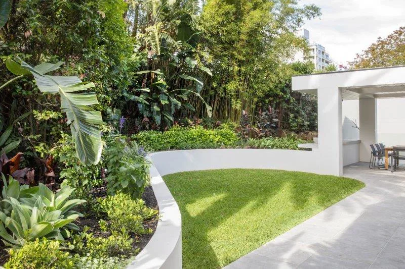Modern outdoor patio with lush garden, green lawn, and white curved wall, with a dining area in the background.