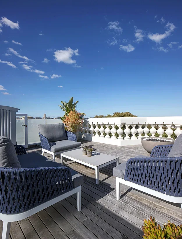 Rooftop terrace with outdoor seating, white coffee table, potted plants, and a decorative white railing under a blue sky with a few clouds.