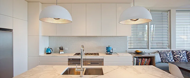 Modern white kitchen with marble island, hanging pendant lights, built-in oven, and window with blinds.