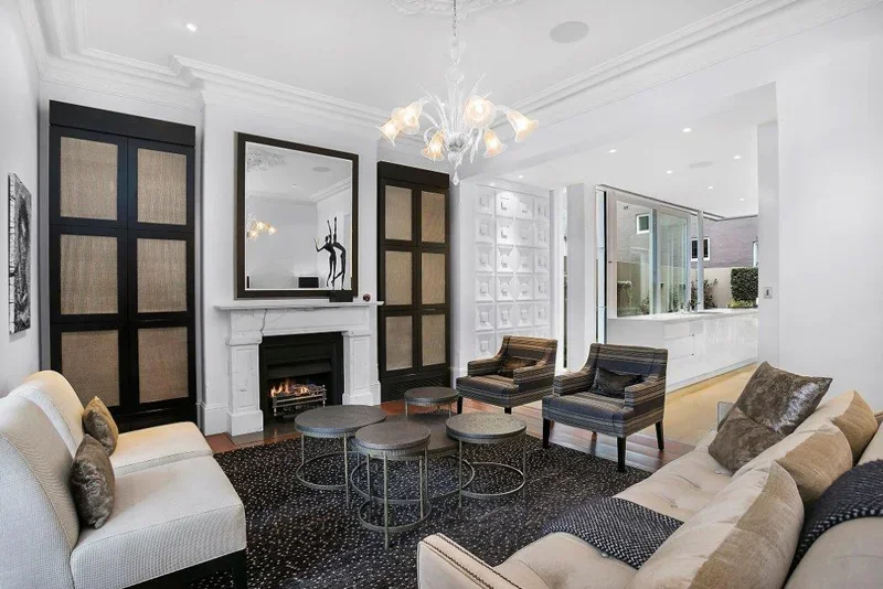 Living room with white walls, a white fireplace with a mirror above it, black built-in cabinets with woven panels, a beige sofa, two striped armchairs, and a modern chandelier.