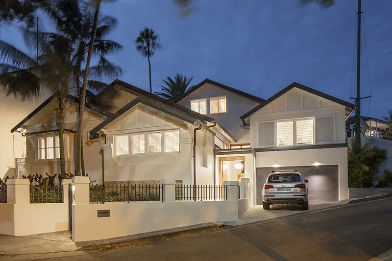 A modern two-story house illuminated at dusk, with a white exterior, large windows, palm trees in the background, and a car parked in the driveway.