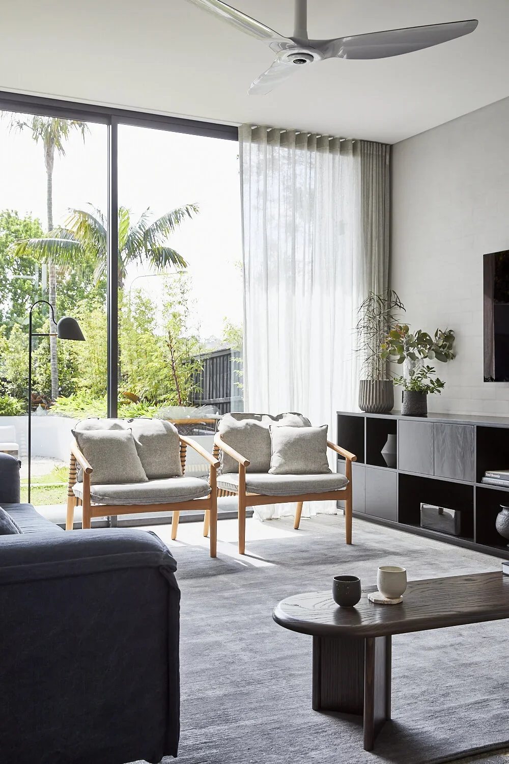 Bright living room with large window, two wooden armchairs with cushions, a black sofa, a modern wooden coffee table with cups, a black and gray console with decorative vases and plants, sheer white curtains, and view of lush greenery outside.
