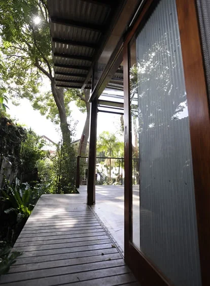 View of a wooden porch with glass sliding doors, surrounded by trees and greenery, under a metal roof with sunlight filtering through.