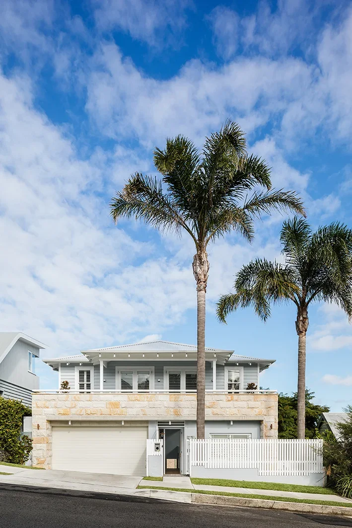 A modern two-story house with a stone facade and a white fence, topped with two tall palm trees, underneath a partly cloudy blue sky.