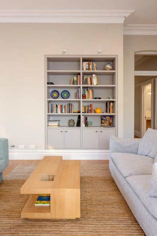 A living room with a built-in wall bookshelf filled with books and decorative items, a light gray sofa on the right, a wooden coffee table in the center, and part of a green armchair on the left, with a beige rug on the floor.