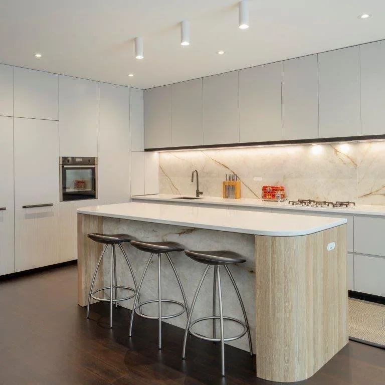 Modern kitchen with white cabinetry, marble backsplash, island with wooden sides, black bar stools, and built-in oven.