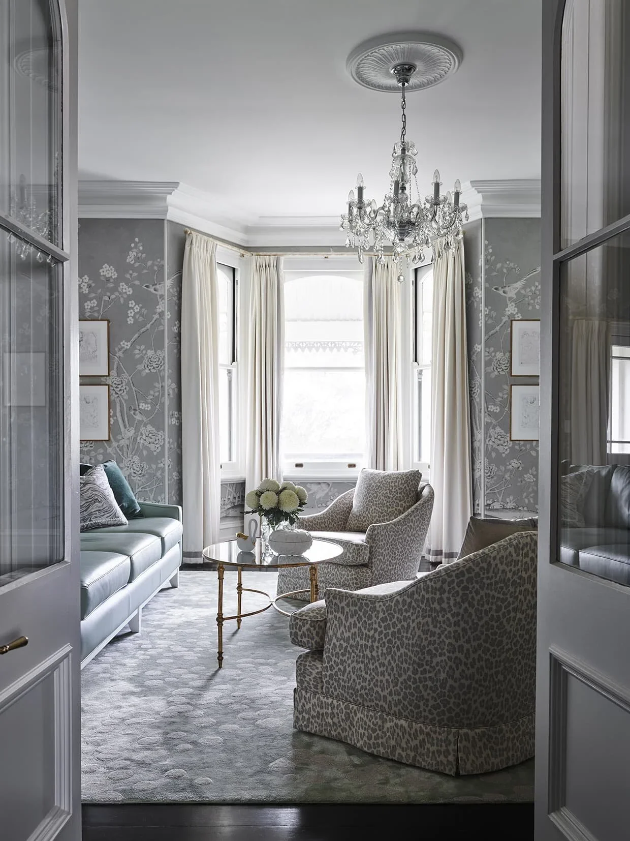 Elegant living room with a chandelier, patterned wallpaper, white curtains, a gray sofa with pillows, two armchairs with animal print fabric, a round glass coffee table with a flower arrangement, and framed artwork on the walls.