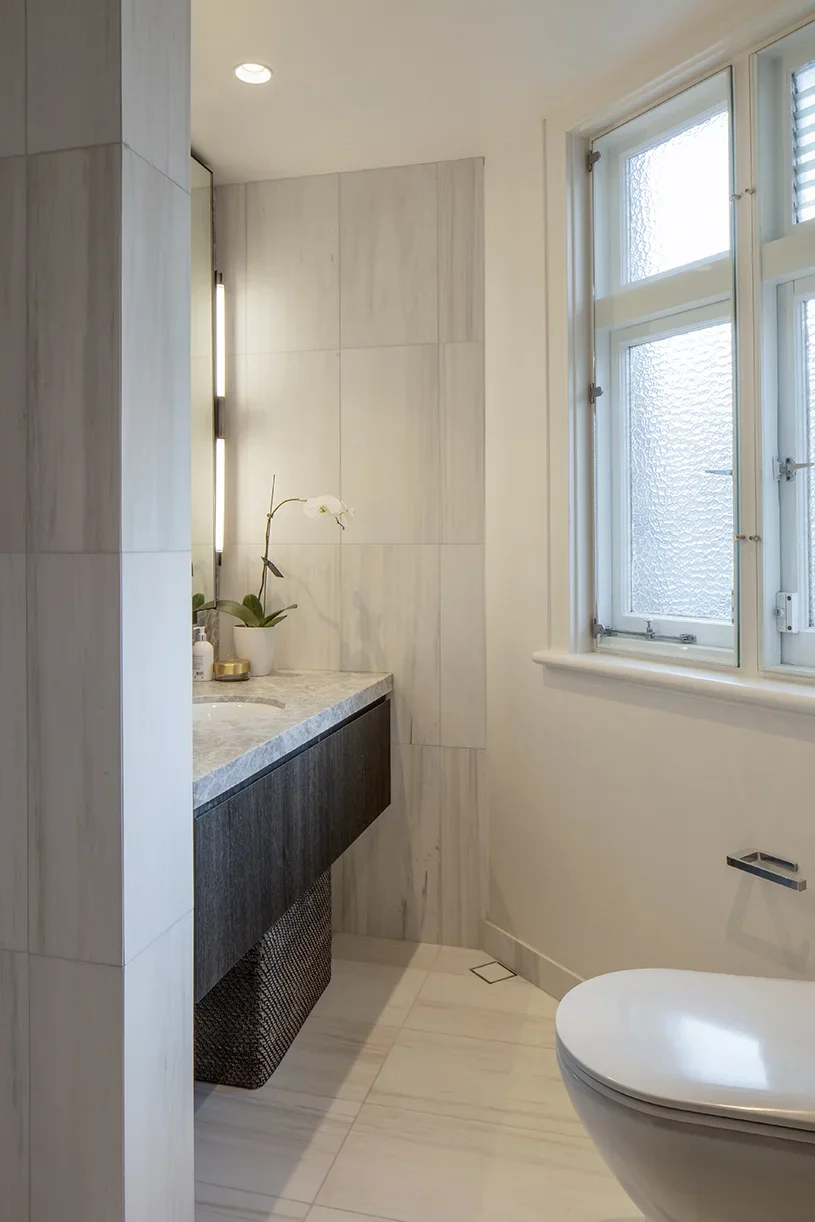 Modern bathroom with a window, a beige tiled wall, a dark wooden vanity with a marble countertop, a white orchid plant, and a wall-mounted toilet.