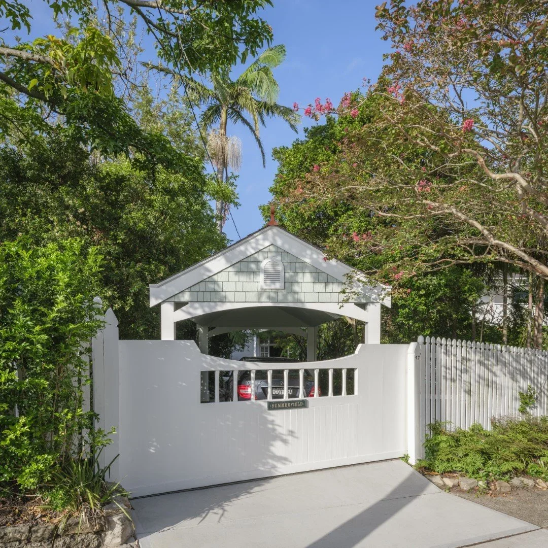White gate and carport leading to a house surrounded by green trees and blooming pink flowers, with palm trees visible in the background and blue sky overhead.