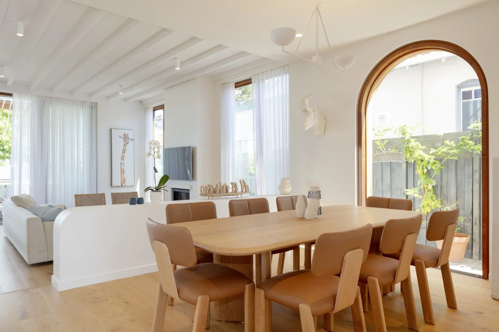 Bright open-concept living and dining area with wooden flooring, tan dining chairs, white walls, large arched window, and minimalistic decor.